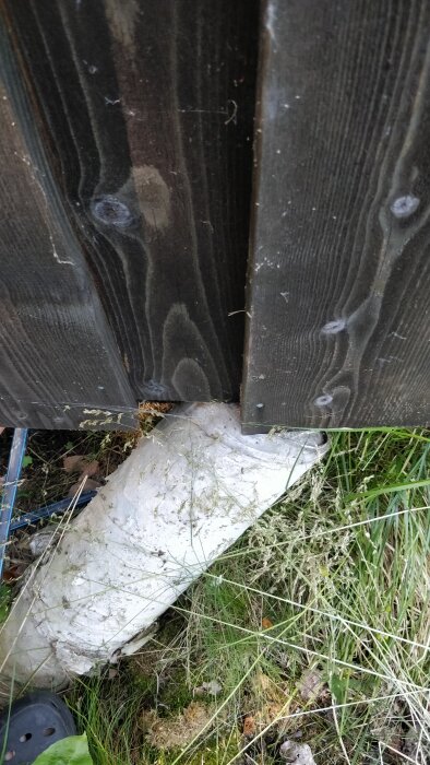 Betongstolpe stöttar en träpanel men har lutat sig på grund av instabil mark. Gräs och mossa syns runt stolpen.