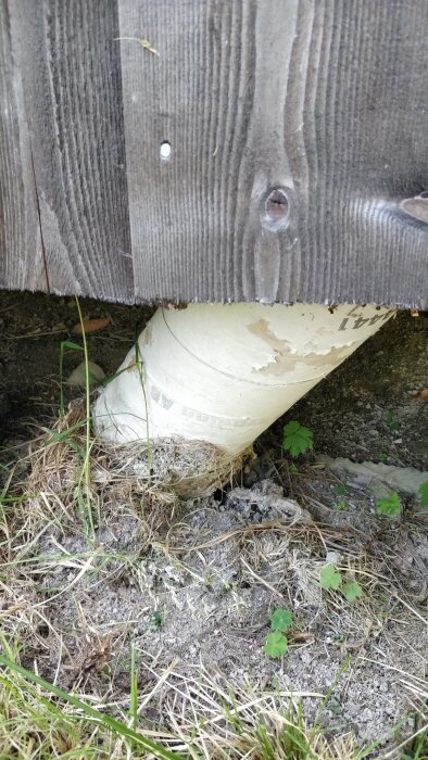 Betongstolpe som lutar mot en trävägg på ett uthus, omgiven av jord och gräs. Stolpen verkar ha vikt sig på grund av lerig mark.