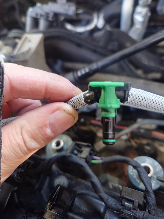 Närbild av en hand som håller i en adapter med grönt fäste, installerat på en bilmotor, omgiven av kablar och kopplingar.