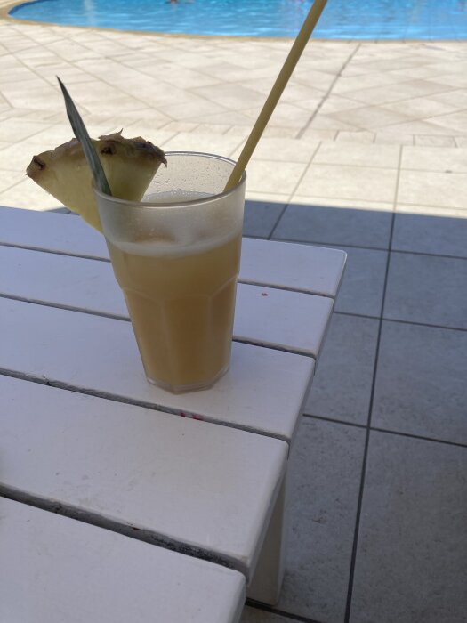 En pina colada med ananasskiva och sugrör på ett vitt bord vid en pool.