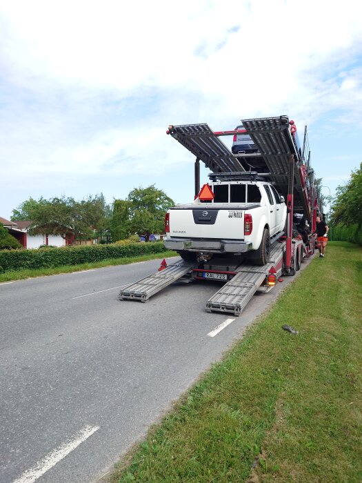 Bil på biltransportsläp parkerad på vägkant, klar för transport eller leverans. Föreställer en vit Nissan Navara lastad för avfärd.