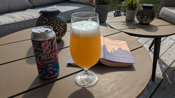 En ölburk från Potum Brewery, ett glas öl och en bok på ett utebord med soffor i bakgrunden.