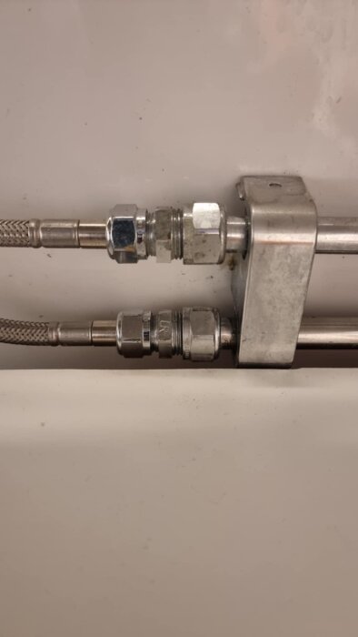 Två flexibla slangar med metallkopplingar och en klämring, anslutna till en metallblock under diskbänk.