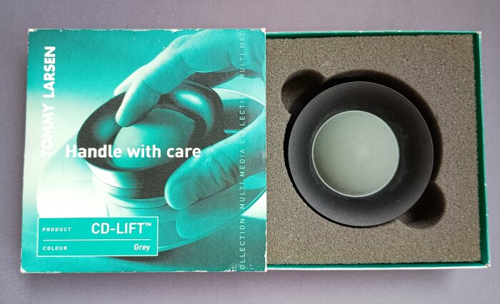 Förpackning med texten "Handle with care" och ett verktyg för att hantera CD-skivor, märkt "CD-LIFT™, Grey", i en vadderad låda.
