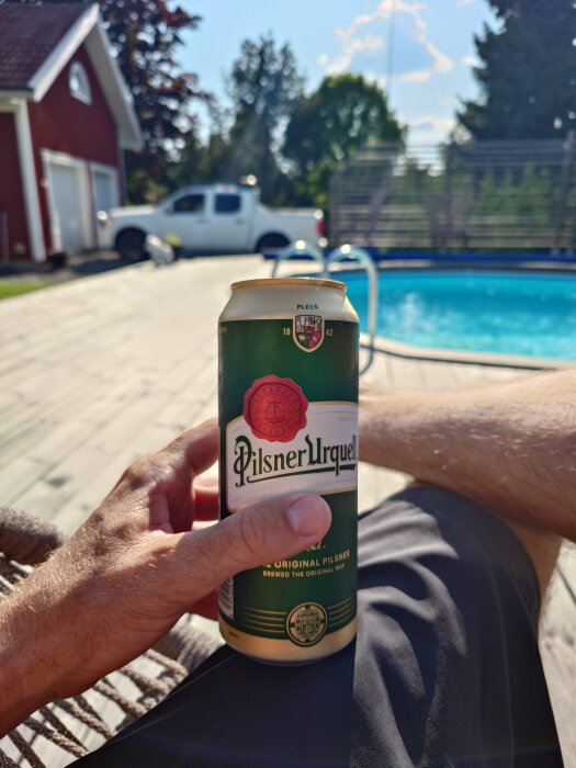 Person håller en burk Pilsner Urquell vid en pool med en vit bil och ett rött hus i bakgrunden, soligt väder.