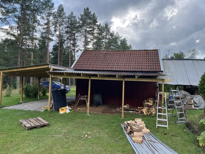Ett trähus med rödfärgad vägg och tegeltak, byggmaterial och stegar på gräsmattan, och en carport med en parkerad blå bil. Skog och grå himmel i bakgrunden.