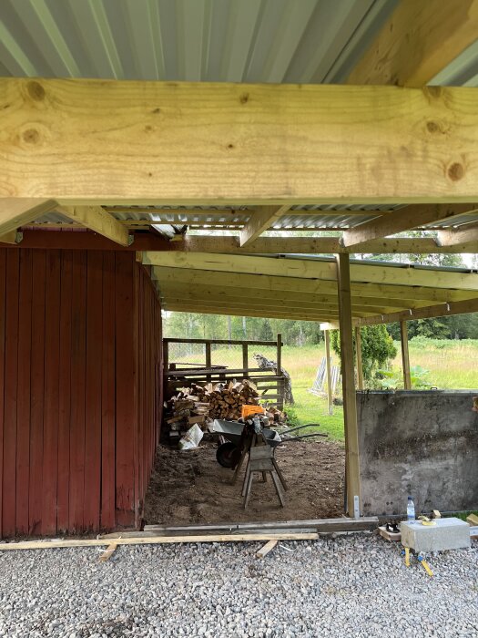 Carport under byggnation med träbjälkar och ett korrugerat tak, ved staplad i bakgrunden och en grönskande omgivning.