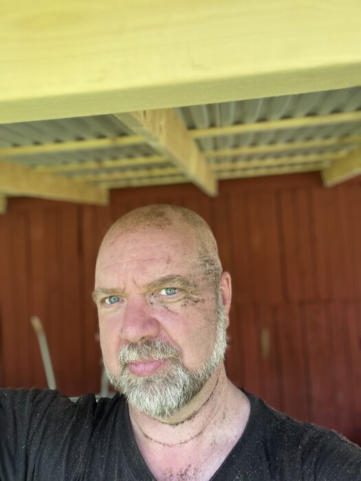 En man med smutsigt ansikte och skägg poserar under ett tak av trä. Bakgrunden är en röd trävägg. Perfekt arbetsväder beskrivs i kontexten.