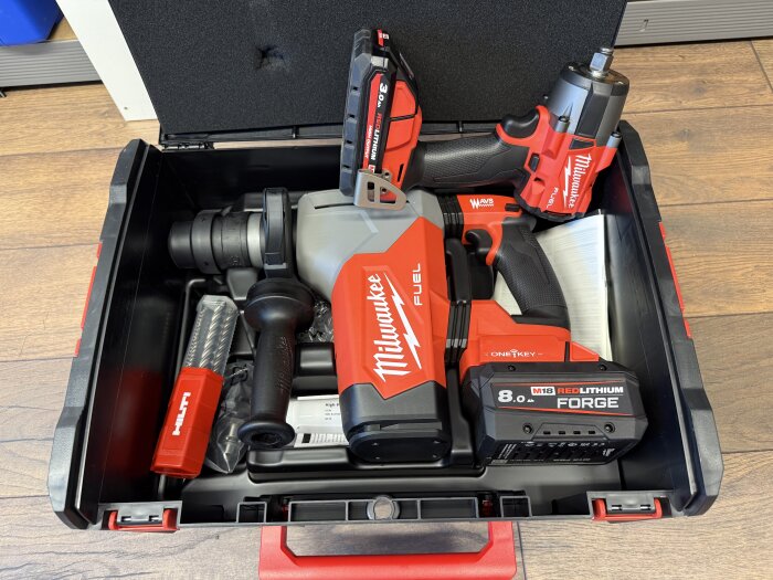 En öppen verktygslåda med röda Milwaukee-verktyg, inklusive en borrhammare och en bultdragare, placerad på ett trägolv.