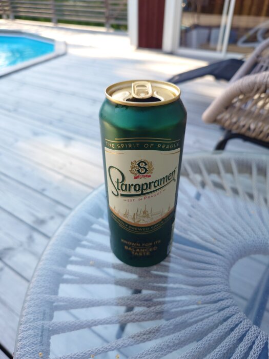 Öppnad burk Staropramen-öl på ett glasbord, med en pool och solstol i bakgrunden. Perfekt för avkoppling under semestern.