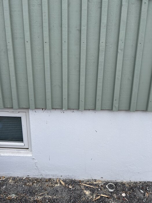 Grön ytterpanel slutar några centimeter ovanför vit grund medan en fönsterkarm syns till vänster. Under ytterpanelen syns råsponten kant i kant.