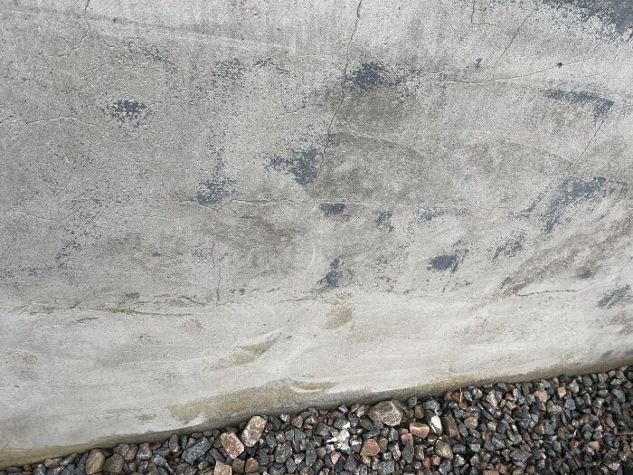 Bilden visar en betongvägg med sprickor och slitage, tillsammans med grus vid basen.