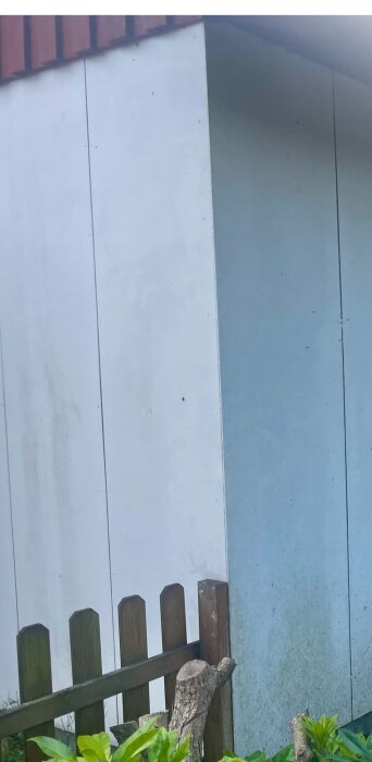 Vitt fasadmaterial misstänkt som eternitskivor på en byggnad vid ett staket och gröna buskar.