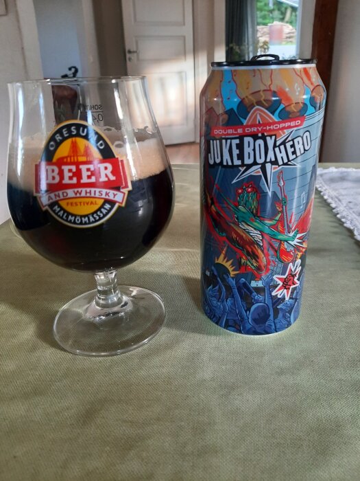Mörk dubbel IPA serverad i ett glas med "Öresund Beer and Whisky Festival" logotyp bredvid en burk "Juke Box Hero" på ett bord.