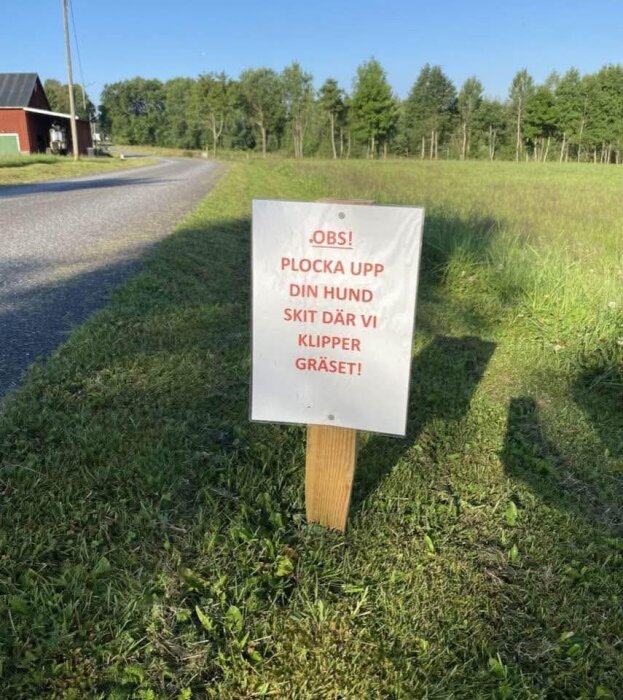 Skylt vid landsväg med texten "OBS! Plocka upp din hundskit där vi klipper gräset!" omgiven av gräs och skog.