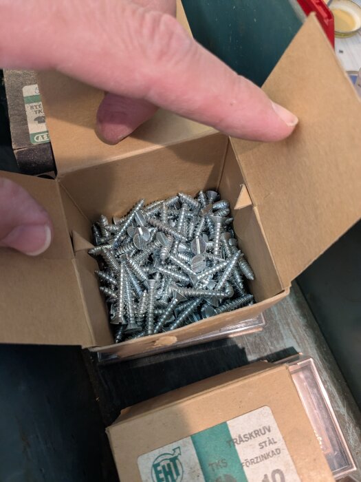 En kartong fylld med flera metallskruvar, betraktares finger pekar in i kartongen.