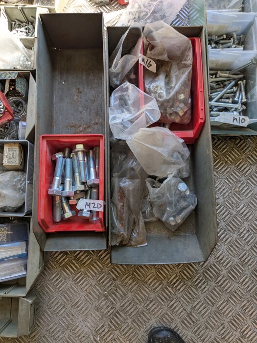 Sortiment av skruv och bult i olika storlekar, M8 till M20, samt spik i lådor och plastpåsar på ett golv med metallmönster.