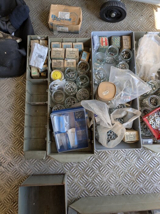 Sorterade behållare med olika storlekar och typer av skruvar och bultar på ett rutigt metallgolv.