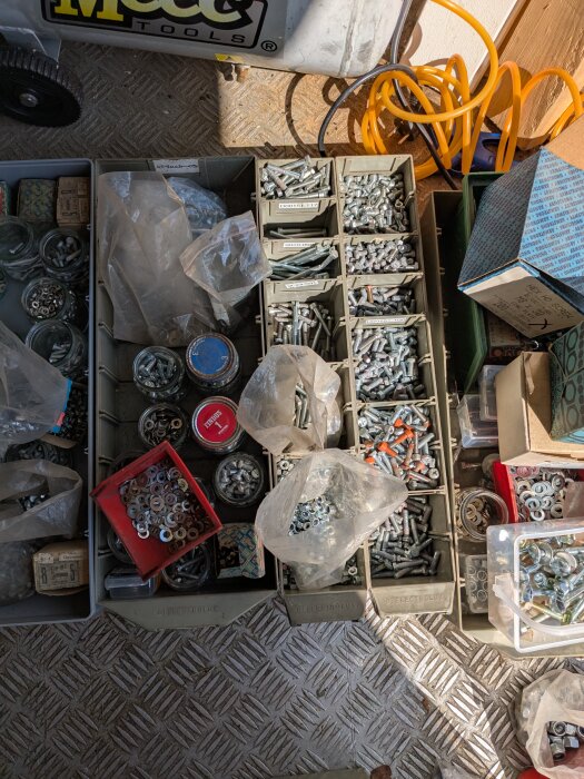 Lådor med skruvar, bultar och spikar i olika storlekar, tillsammans med plastpåsar och burkar på ett golv.