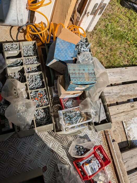 Lådor med diverse skruvar, muttrar och spikar i varierande storlekar, placerade utomhus på en träyta med gräs bredvid.
