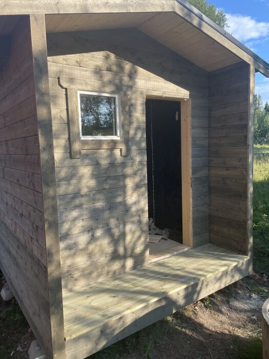 Isolerad stuga med träpanel och litet fönster, dörrområde klart. Soligt bygge omgiven av träd, paus i projekt för semester till norr och grannland.