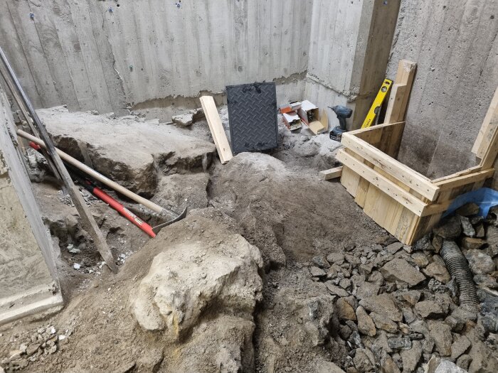 Byggarbetsplats med schakt och betongvägg. Verktyg och material är utspridda, inklusive en träform, en borrmaskin och en nivå.