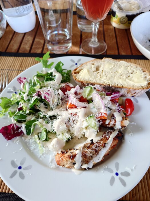 Tallrik med grillad kyckling, sallad med tomat, gurka, fetaost, parmesan och Caesar-dressing. Bröd med smör och dryck i bakgrunden.