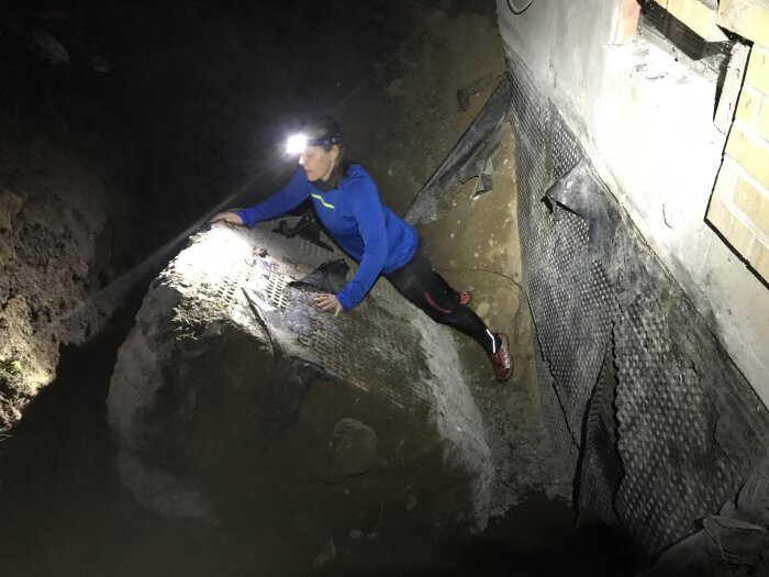 Person med pannlampa undersöker en stor betongklack i en grop intill en husvägg, omgiven av jord och plastisolering.