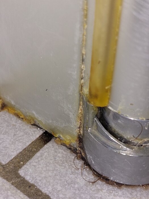 Närbild av en spricka i kakel och mögelbildning i ett badrum nära en duschkabin och golv med fogmassa.