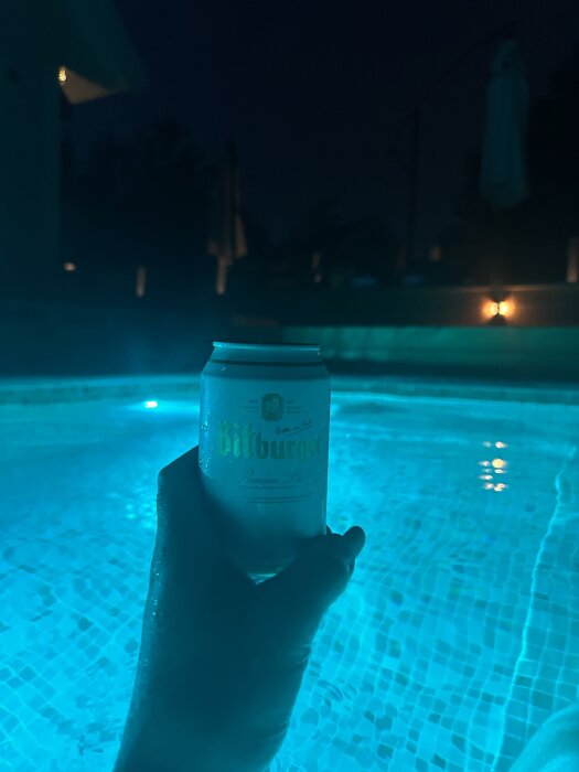 En hand håller en burk Bitburger-öl över en upplyst swimmingpool på natten.