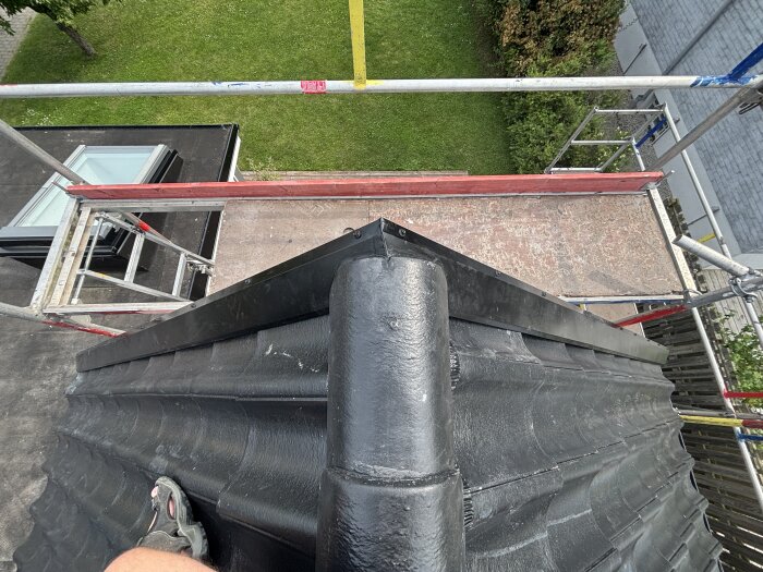 Person står på ett svart tegeltak med byggställning nära, tittar mot besiktade vindskivor och en gräsmatta.