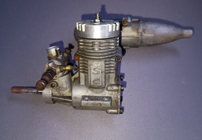 Gammal modellflygplansmotor med SC-logotyp, kylflänsar och bränslenipplar, används för glödstiftmotorer med oljeblandad metanol.