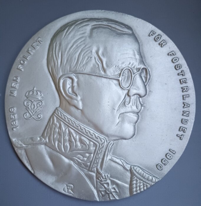 Relief av man i profil med glasögon, militär uniform, och text "Med folket 1938" och "För fosterlandet 1950" ingraverat på kanten.