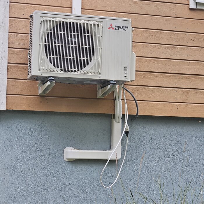 Luftvärmepump på träfasad med synlig elkontakt hängande.