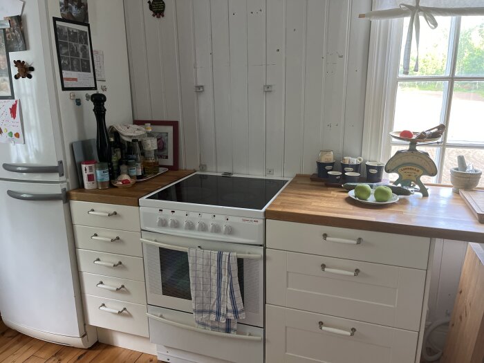Kök med vit spis mot trävägg, kylskåp till vänster, träbänkskiva till höger med äpplen och köksvåg, diskussion om installation av ny bänkskiva.