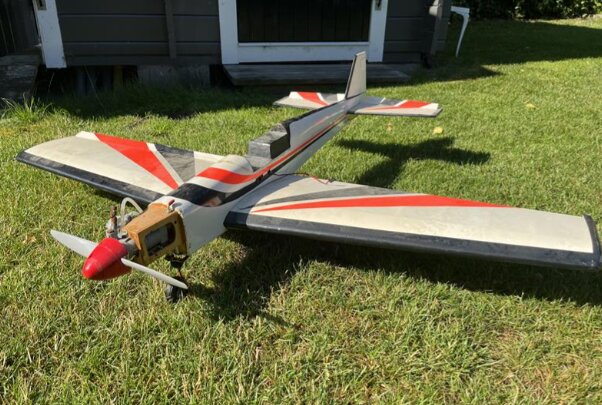 Radiostyrt flygplan med röda och svarta markeringar på gräsmatta framför en friggebod.
