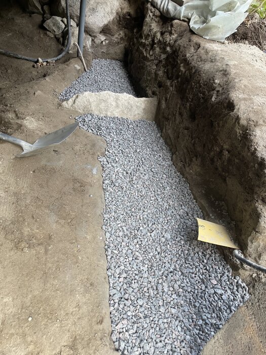 Grusfylld ränna invid jordvägg, med verktyg och material synliga, del av pågående byggprojekt.