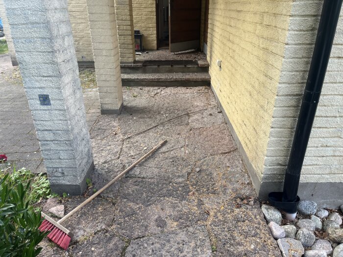 Husets hörn med gul tegelvägg, svart stuprör, och ett område med stenplattor. En kvast ligger på marken bredvid en stenpelare.