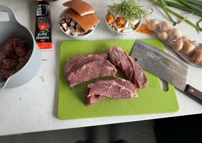 Köttskivor på en grön skärbräda med kniv, bredvid morötter, lök och örter i skål. Tomatpuré och fläsk i bakgrunden.