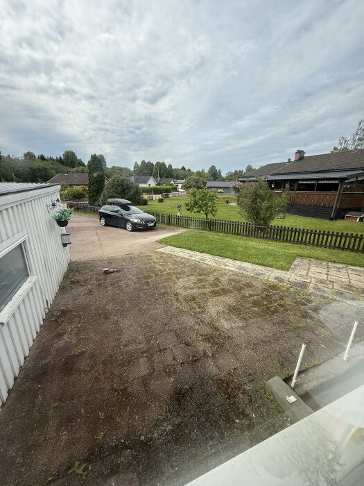 Vy över en gårdsplan med en svart bil parkerad, ett grönområde och en kapad linje i asfalten för framtida grusläggning.