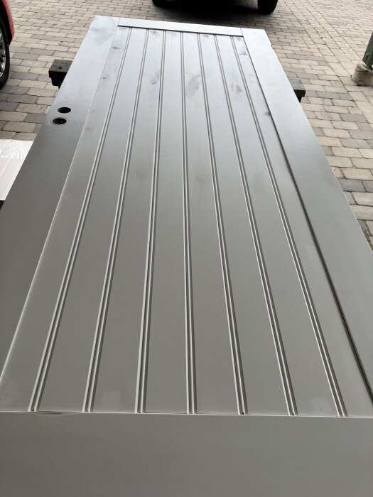 Nymålad grå dörr med vertikala paneler, placerad på en stenbelagd uppfart, redo för montering.