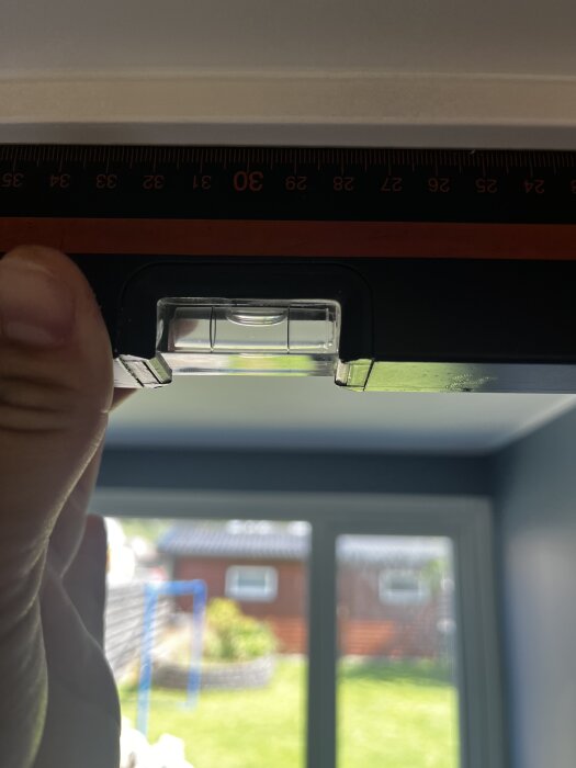 Hand håller vattenpass mot överstycke som inte är i våg, med fönster och trädgård i bakgrunden.