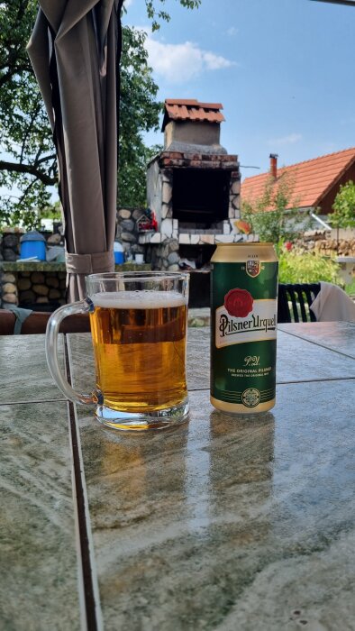 Ölglas och burk av Pilsner Urquell på ett bord med stenugn och trädgård i bakgrunden.