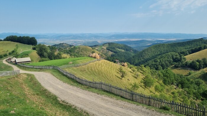 Landskap med grusväg, gröna kullar och bondgård, sedd från hög höjd med utsikt över avlägsna berg.