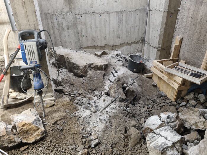 Byggarbetsplats med borrmaskin och jordhög invid betongvägg, omgiven av stenar och träpallar med metallrör.