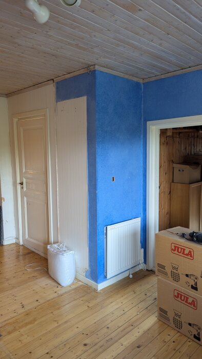 Hörn av ett rum med blå vägg, vita lister och dörrkarmar, en radiator, trägolv, och kartonger märkt "Jula.