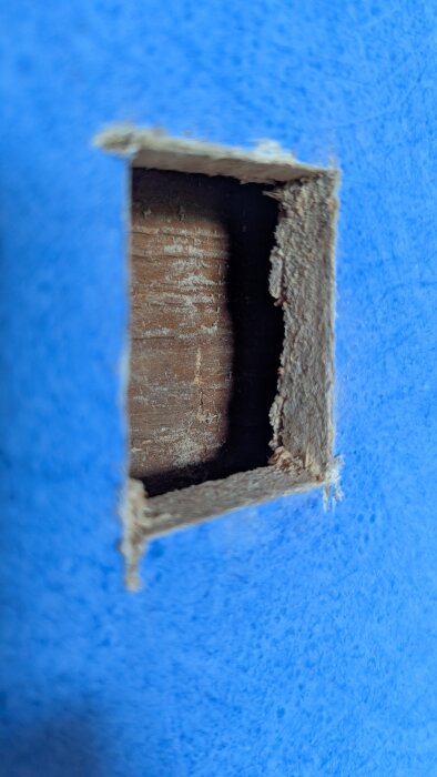 Utskuret fyrkantigt hål i väggen visar en trädstruktur bakom blå yta, används för att bedöma möjligheten att ta bort ett hörn i ett timmerhus.