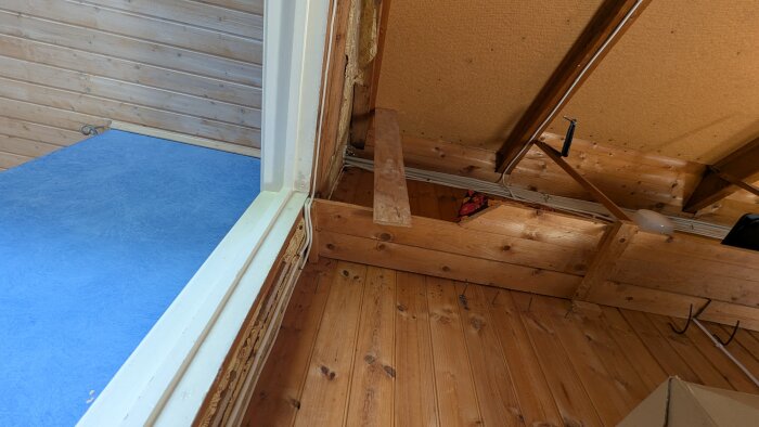 Ett hörn av ett timmerhus med trätak, vägg och golv synliga. Strukturen ser robust ut med exponerade balkar och blått golv.