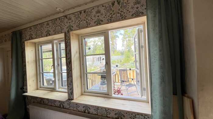 Två fönster med skräddarsydda fönstersmygar i trä, tapetserade väggar och utsikt mot en solig uteplats med stolar och träd.