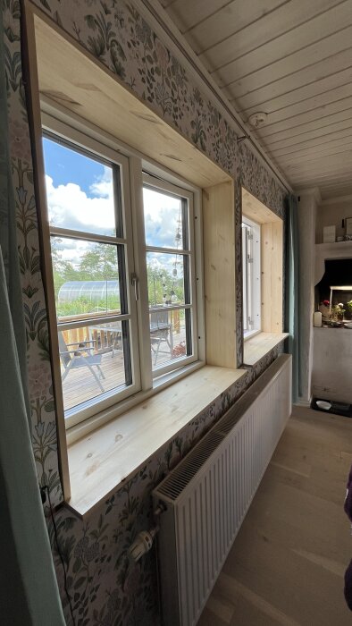 Fönstersalning i ljusa träpaneler med blommig tapet runt och utsikt mot en altan med bord och stolar.